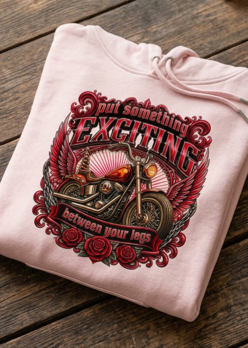Front of the pink Put Something Exciting premium hoodie with the Put Something Exciting motorcycle graphic, laid flat on a wooden table